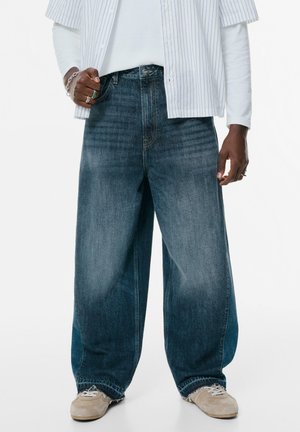 Jeans en denim à jambe large de couleur bleu foncé délavé avec une coupe ample. Les ourlets effilochés et les légères marbrures ajoutent de la texture. Portés avec des baskets beiges.