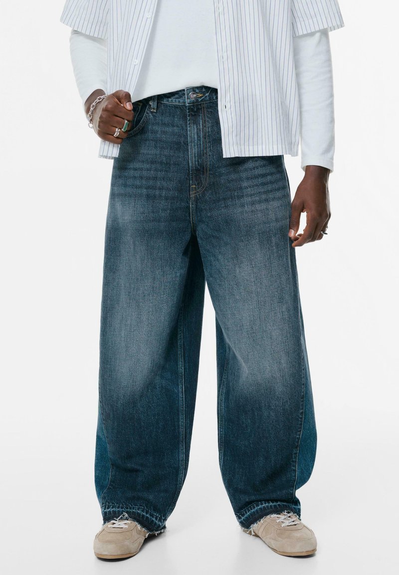 Široke traperice u izblijedjeloj tamnoplavoj boji s opuštenim krojem. Iznad ruba su izribane, a suptilno bora dodaju teksturu. Nošene s bež tenisicama.