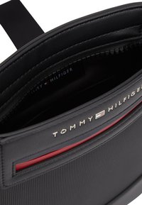 Μαύρη τσάντα χιαστί με υφή, με κόκκινη λεπτομέρεια, χρυσό λογότυπο Tommy Hilfiger και ορατή εσωτερική τσέπη με φερμουάρ και ετικέτα του brand.
