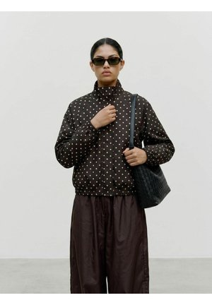 Femme portant une veste noire à pois, un pantalon large foncé, des lunettes de soleil, tenant un sac à bandoulière noir texturé, debout devant un fond uni.