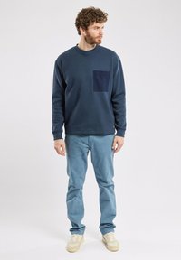 Sweatshirt marine avec une poche à rabat, associé à un pantalon bleu clair et des baskets beiges ; présente un col rond et une coupe décontractée.