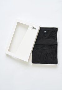 Sort t-shirt og matchende bukser, foldet inde i en hvid kasse med en klar vindue. Kassen har et låg og logoet "Jbs" på siden.