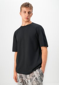 Tânăr îmbrăcat cu un tricou simplu negru cu mânecă scurtă și pantaloni scurți cu un pattern de frunze, stând pe un fundal alb.