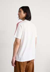 T-shirt de algodão branco com riscas vermelhas nos ombros, mangas curtas e decote redondo, combinado com calças castanhas.