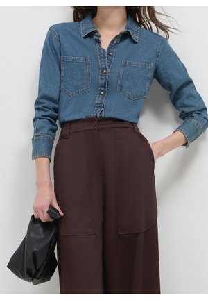 Vrouw die een blauw denimshirt draagt, in een hoge bruine broek gestopt, met een kleine zwarte handtas en één hand in haar zak.