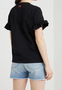 Camiseta de algodón negra con mangas cortas y con volante, y un pequeño logo rojo en la parte de atrás; combinada con shorts de mezclilla azul claro.