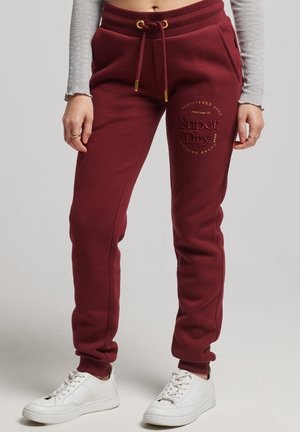 Person iført bordeaux SuperDry joggingbukser med snøre i taljen, kombineret med hvide sneakers og en cropped grå langærmet top.