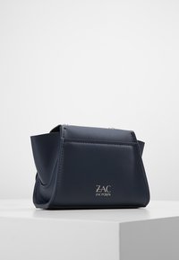 Mörkblå läderhandväska från Zac Posen med strukturerade sidor, silverlogotyp på framsidan och en metallkedjerem som syns vid toppkanterna.