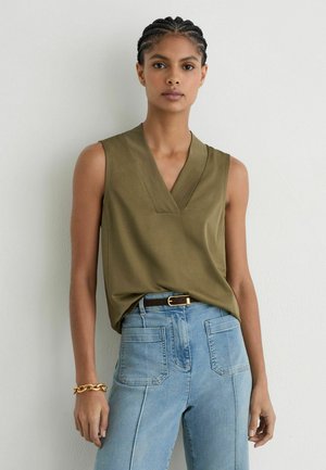 Kvinde med fletninger iført en olivengrøn ærmeløs V-hals top, lyseblå jeans med høj talje og et guldarmbånd, stående mod en hvid væg.