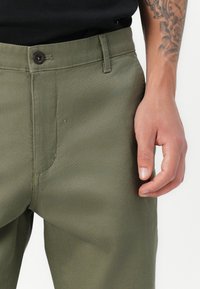 Khaki green trousers with a smooth texture, featuring a button closure and belt loops. A hand is visible near the waistband.