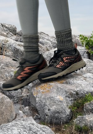 Persona con leggings grigi e calzini spessi grigi che cammina su terreno roccioso con scarpe da escursionismo nere e arancioni.