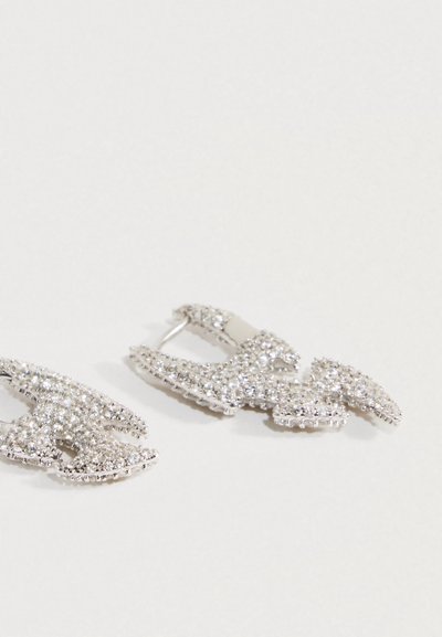 Par srebrno toniranih uhanov v obliki zaponk, okrašenih z majhnimi prozornimi kristalčki, na gladki beli površini.