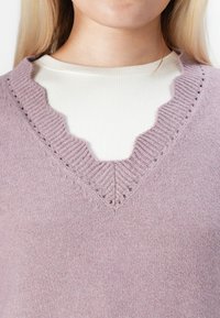 Close-up de uma pessoa a usar uma camisa branca de malha sob um suéter de malha lavanda com um colarinho V profundo em ondas.