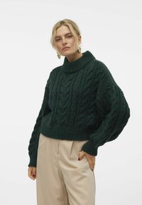 Donkergroene kabelgebreide trui met hoge hals, geribbelde manchetten en een cropped ontwerp, gecombineerd met losse beige broeken.