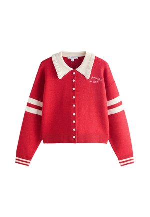 Ung kvinde med langt blondt hår iført store mørke solbriller og en rød cardigan med hvide striber og en hvid krave står udenfor.
