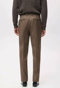 Brązowe, dopasowane spodnie o gładkiej fakturze, z dwoma tylniami kieszeniami zapinanymi na guziki oraz prostymi nogawkami. Noszone z dzianinowym swetrem.