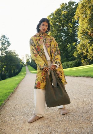 Junge Frau steht auf einem Gartenweg, trägt einen gelben Mantel mit Blumenmuster, helle Hose, hält eine große dunkle Handtasche, mit Bäumen im Hintergrund.