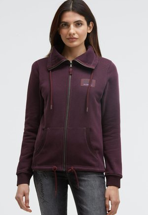 Purple zip-up hoodie with a high collar, drawstring waist, two front pockets, and a fabric patch logo on the chest. Smooth texture.