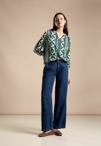 Blusa verde e bianca a fantasia con maniche lunghe, abbinata a jeans a vita alta blu scuro a gamba larga e scarpe flat marroni. Sfondo semplice.