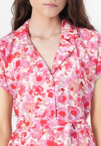 Blomstret kjole med et pink og rødt akvarelmønster, korte ærmer, krave, frontknaplukning og en(bundle) der afrunder taljen.