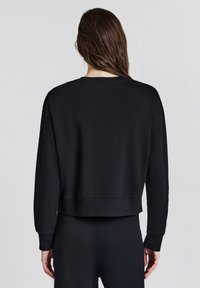 Donna con lunghi capelli castani che indossa una semplice felpa nera a maniche lunghe e pantaloni neri, vista di spalle su uno sfondo neutro.