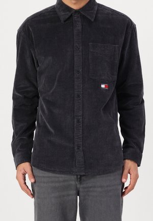 Homme portant une chemise en velours côtelé noire à boutons avec une poche poitrine et un petit logo Tommy Jeans, associée à un pantalon gris foncé.
