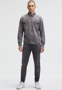 Graues Denimhemd mit langen Ärmeln, Knopfleiste, zwei Brusttaschen und kontrastierenden Nähten, kombiniert mit dunklen Jeans und weißen Sneakern.