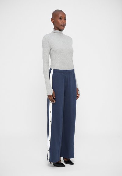 Vrouw in een grijze langdouch turtleneck en marineblauwe wijde broek met witte zijstreep en knopen, staand tegen een witte achtergrond.