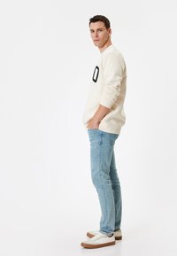 Beige crewneck sweatshirt med svart nummerdetalj, ljusblå jeans och vita sneakers med bruna accenter. Modellen står avslappnat.