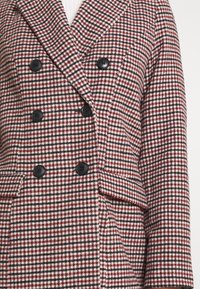 Blazer de doble botonadura en un patrón de piel de gallo rojo, negro y blanco; cuenta con botones negros y una tela texturizada con un corte entallado.