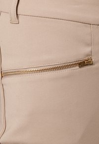 Beige-kankaiset housut, joissa on vaakasuora kultainen vetoketjullinen tasku ja vyölenkki näkyvissä vasemmassa ylhäisessä kulmassa.