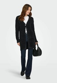 Cardigan noir en tissu pelucheux avec manches longues, col en V, boutons sur le devant, deux poches, associé à un jean bleu et un sac à main noir.