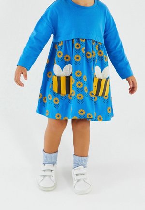 Un tout-petit portant une robe bleue avec des fleurs jaunes et des poches à thème abeille, associée à des chaussettes bleu clair et des chaussures blanches à Velcro sur fond blanc.
