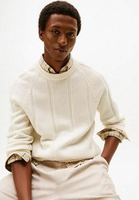 Maglione crema in maglia con trama a coste e righe verticali, indossato sopra una camicia leggera a quadri. Il modello porta pantaloni beige.