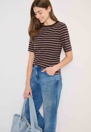 Jeune femme souriante portant un t-shirt rayé marron et rose ainsi qu'un jean bleu, tenant un sac fourre-tout en denim bleu clair d'une main.