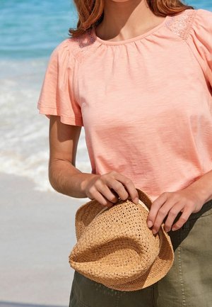 Jasnoróżowa bluzka z krótkim rękawem, zdobiona marszczonym dekoltem i koronkowymi akcentami. Kobieta trzyma pleciony kapelusz słomkowy w tle plaży.