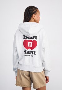 Grauer Kapuzenpullover mit einem großen roten Herz und schwarzem Text auf dem Rücken. Weiches Material, lockere Passform, gerippte Bündchen und Saum. Kombiniert mit beigen Shorts.