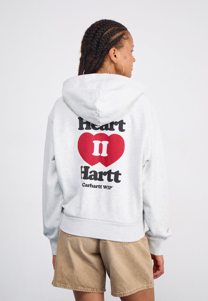 Grauer Kapuzenpullover mit einem großen roten Herz und schwarzem Text auf dem Rücken. Weiches Material, lockere Passform, gerippte Bündchen und Saum. Kombiniert mit beigen Shorts.