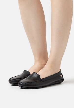 Des pieds chaussés de mocassins en cuir noir, avec des détails brodés et un petit logo métallique sur le côté, sur un fond blanc.