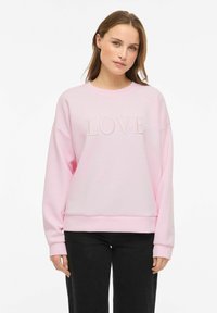 Sweatshirt rose clair avec texte "LOVE" en relief sur le devant, encolure arrondie et épaules tombantes. Associé à un pantalon noir.