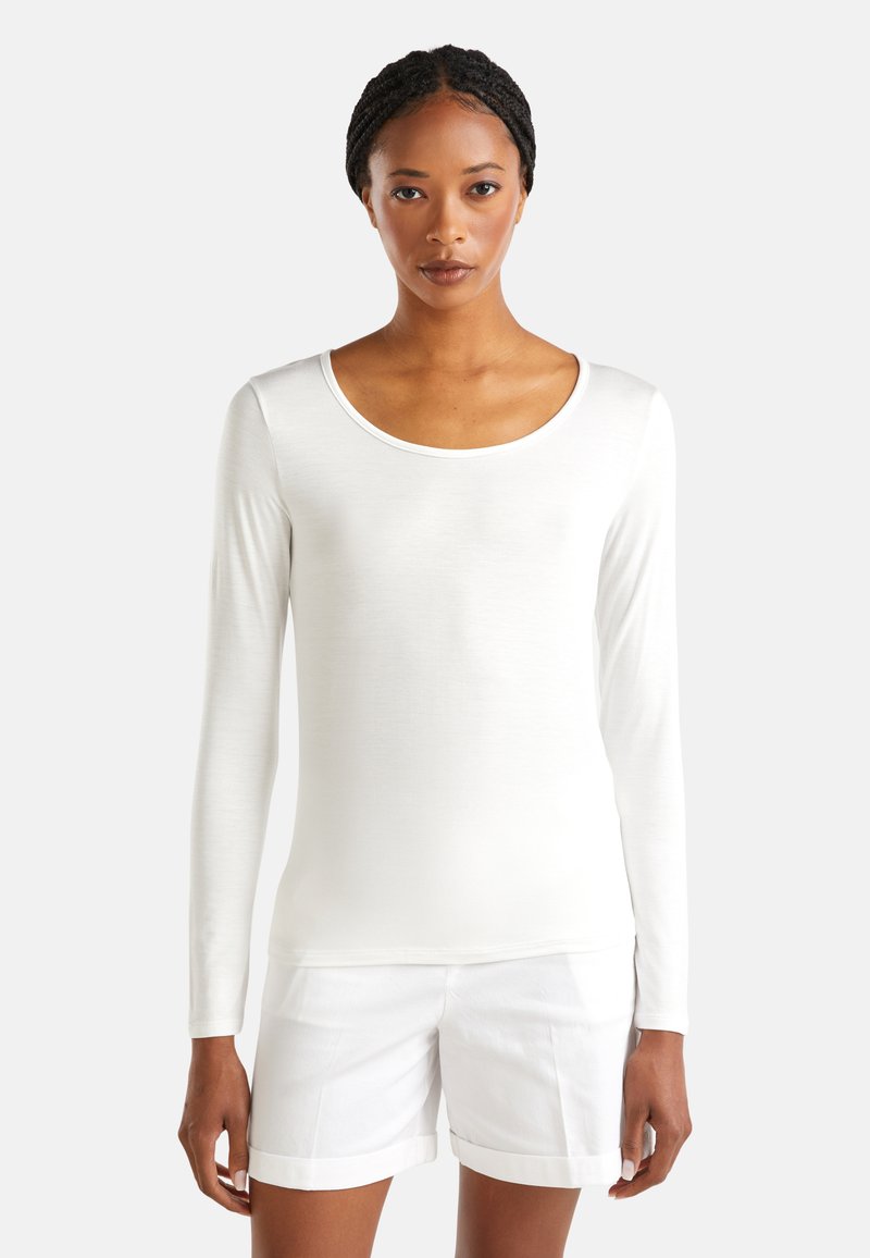 Vrouw met gevlochten haar, gekleed in een witte top met lange mouwen en ronde halslijn, en witte shorts met opgerolde zomen, staand tegen een effen achtergrond.