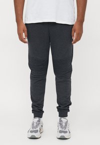 Man wearing dark gray ribbed knee joggers, white sneakers, white socks, and a tucked white T-shirt against a plain background.