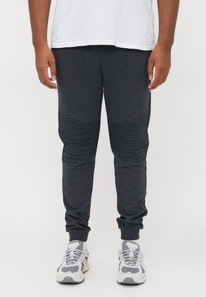 Man wearing dark gray ribbed knee joggers, white sneakers, white socks, and a tucked white T-shirt against a plain background.