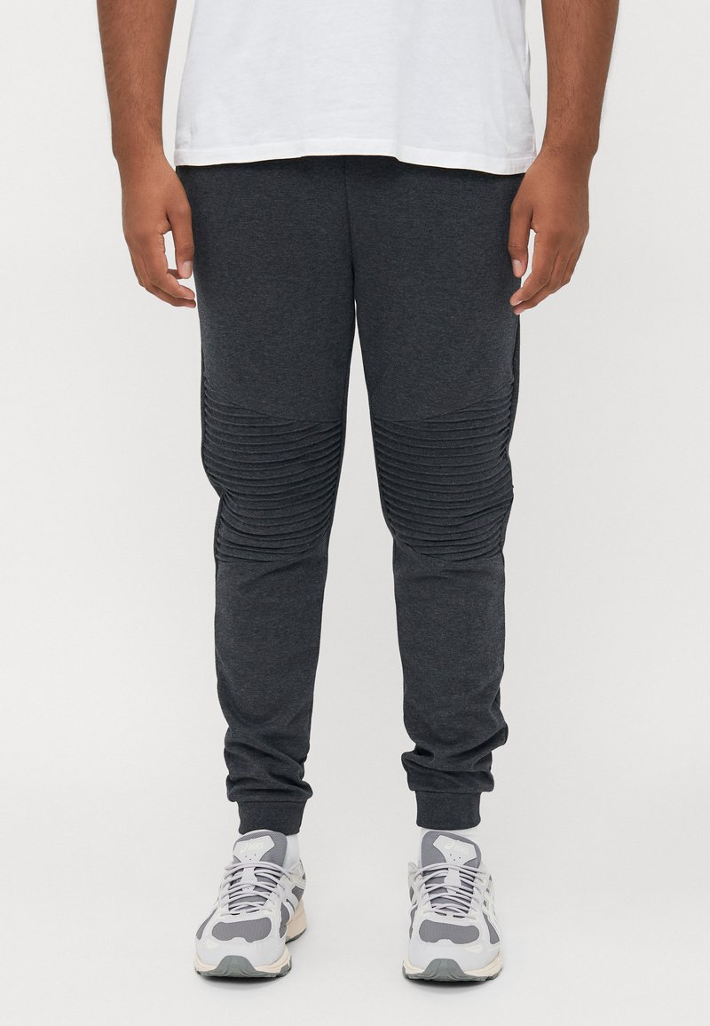 Man wearing dark gray ribbed knee joggers, white sneakers, white socks, and a tucked white T-shirt against a plain background.