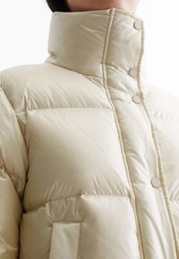 Beige pufferjakke med høy krage, trykknapper og quiltet tekstur; har sidelommer og en glatt, skinnende stofffinish.