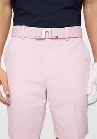 Vaaleanpunaiset shortsit, joissa on teksturoitu kangas, leveä vyö metallisen soljen kanssa ja kaksi sivutaskua. Yhdistettynä tummansiniseen paitaan ja valkoiseen hansikkaaseen.