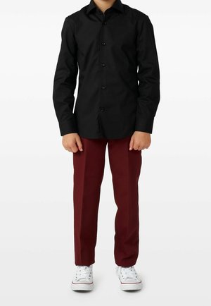 Barn stående iført en sort skjorte med knapper, burgunderbukser og hvide sneakers mod en ensfarvet hvid baggrund.