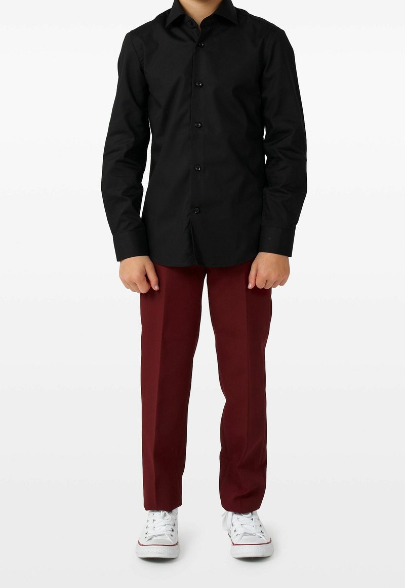 Enfant debout portant une chemise noire à boutons, un pantalon bordeaux et des baskets blanches sur un fond blanc uni.