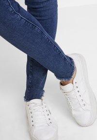 Jeans ajustados de mezclilla azul oscuro con dobladillo deshilachado, combinados con zapatillas de lona blancas que tienen una suela texturizada y costuras visibles.