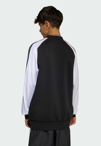 Crna i bijela dukserica s visokim ovratnikom, dugim rukavima i crvenim detaljima. Glatka tkanina i rebrasti manžeti. Klasičan sportski dizajn.